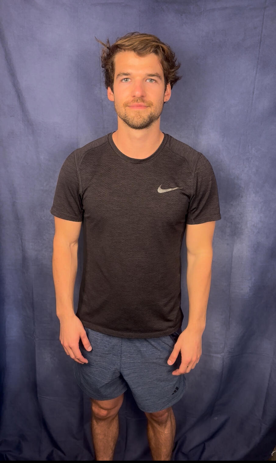 A caucasian man with ear length brown hair, green eyes, and white teeth wearing a black T-shirt stands in front of a blue background for a full body slate shot.