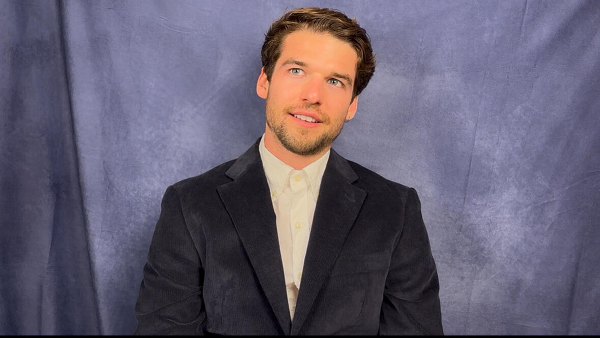 An example of a self tape. A caucasian man stands in front of a blue background with ear length brown hair, green eyes, and white teeth. He is wearing a navy blue sports coat and looks excitedly toward someone standing above him, off-screen.