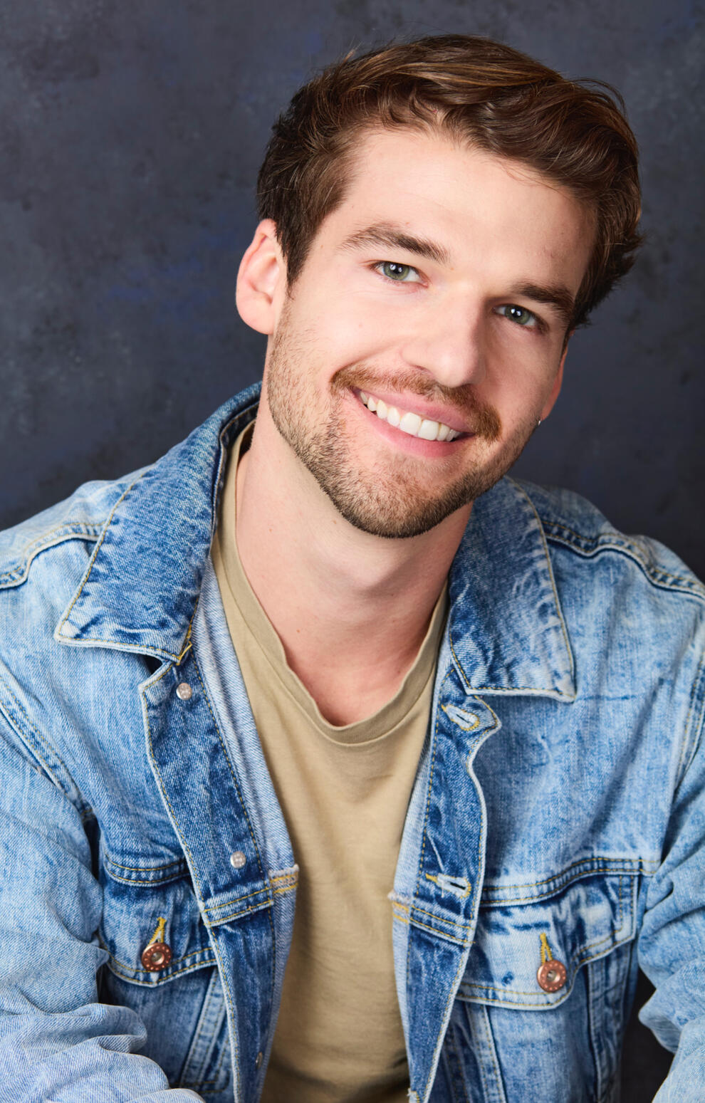 Headshot A headshot of a white man with green eyes and medium length brown hair. He’s wearing a light green T-shirt underneath a blue denim jacket, looking solemnly into camera.