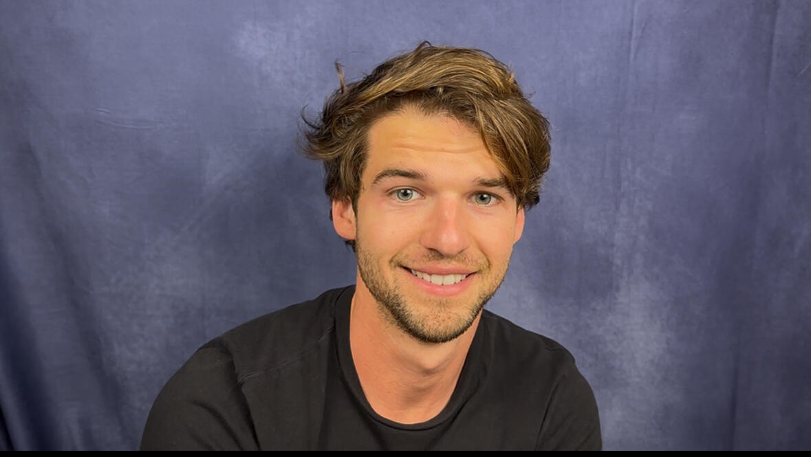 A caucasian man with ear length brown hair, green eyes, and white teeth wearing a black T-shirt smiles awkwardly and looks expectantly as he acts toward someone in front of him, but off screen.